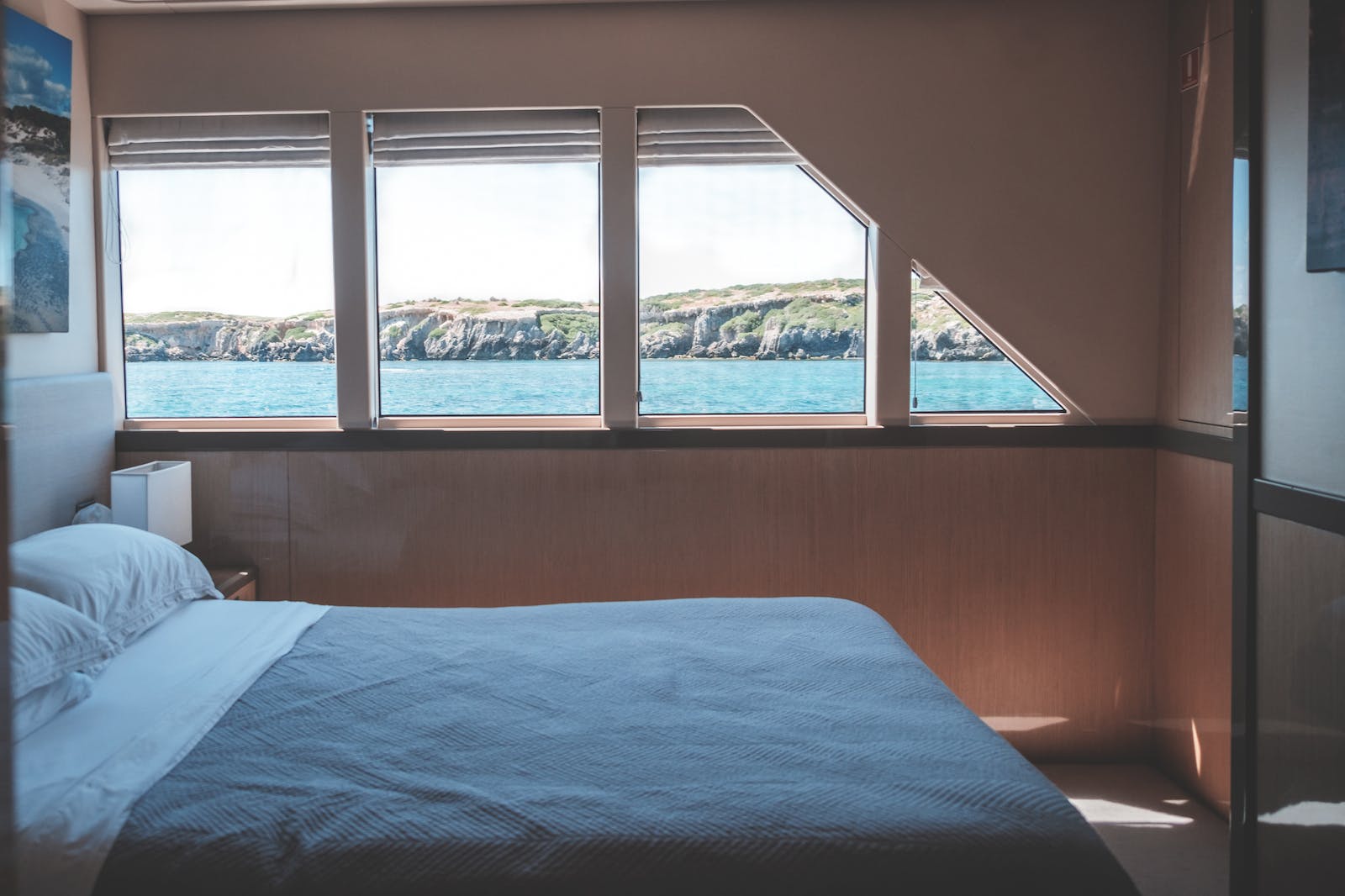 Interior of cabin in modern yacht with comfortable bed and windows overlooking picturesque sea and rocky cliff on sunny day